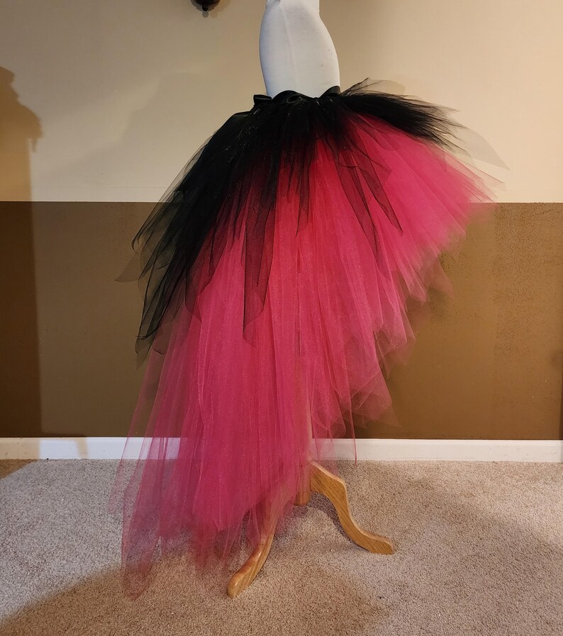 May include: A black and hot pink tulle skirt displayed on a mannequin. The skirt has a high-low design, with black tulle at the top that fades into a vibrant pink. The skirt is gathered at the waist with a black ribbon.