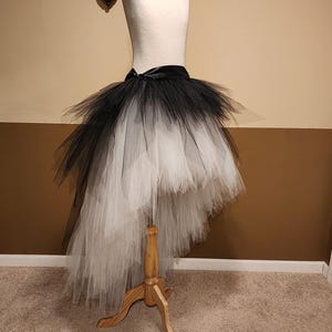 May include: A black and white ombre tutu skirt displayed on a mannequin. The skirt features a high-low design, with a black waistband and a satin bow. The tulle transitions from black at the top to white at the bottom.