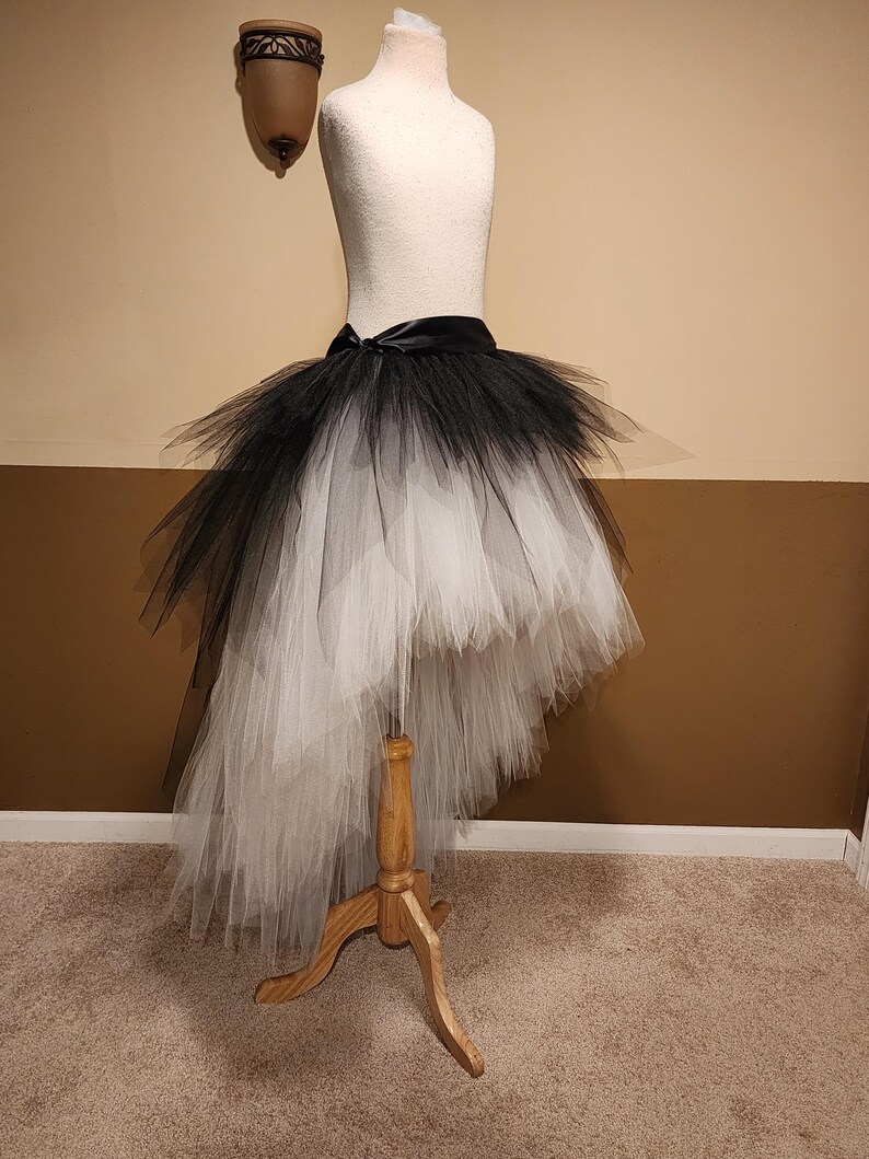 May include: A black and white ombre tutu skirt displayed on a mannequin. The skirt features a high-low design, with a black waistband and a satin bow. The tulle transitions from black at the top to white at the bottom.