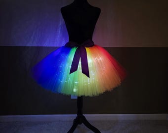 May include: A rainbow-colored tutu skirt with LED lights, displayed on a black mannequin. The skirt transitions through the colors of the rainbow, with a dark purple bow at the waist. The skirt is illuminated, creating a vibrant effect.