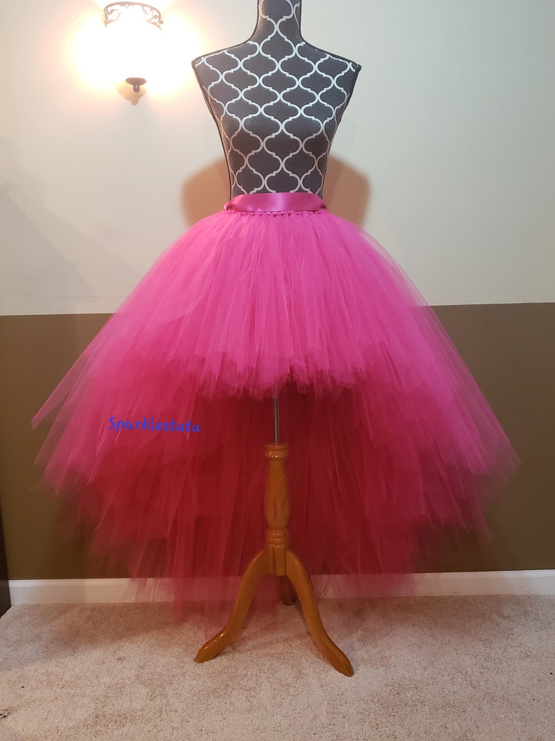 Puede incluir: Una falda de tul de color rosa intenso de corte alto-bajo con una cinta de raso en la cintura. La falda est&aacute; en un maniqu&iacute; y la falda es larga en la parte posterior y corta en la parte delantera.
