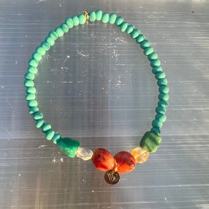 May include: A turquoise beaded necklace with a gold clasp. The necklace features a variety of gemstone beads, including green, red, and clear stones. A small gold charm with a symbol hangs from the necklace.