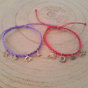 May include: Two handmade bracelets. One is purple, the other is red. Each bracelet has a braided cord and small silver charms. The purple bracelet has a star, whale, planet, and butterfly charm. The red bracelet has a bear, sun, bird, and origami boat charm.