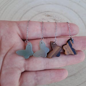 Peut inclure: Quatre pendentifs en forme de papillon suspendus à de fines chaînes en métal argenté. Deux pendentifs sont d'un vert sourd uni, tandis que les deux autres présentent un motif rayé marron et noir. Chaque pendentif est fixé par un anneau.
