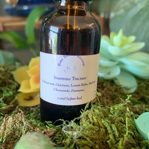 May include: A brown glass bottle with a black cap, labeled "Insomnia Tincture." The label lists ingredients like Valerian root and Chamomile. The bottle sits on a bed of green moss, with yellow and green plants in the background.