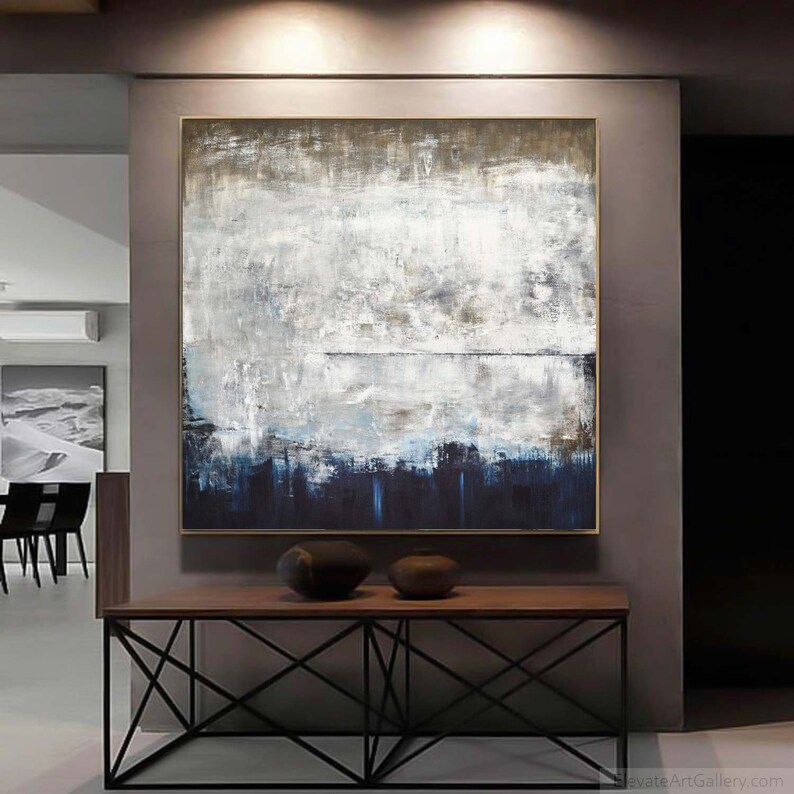May include: Abstract painting featuring shades of white, grey, and blue. The painting is framed and hanging on a wall above a wooden table with a black metal base.