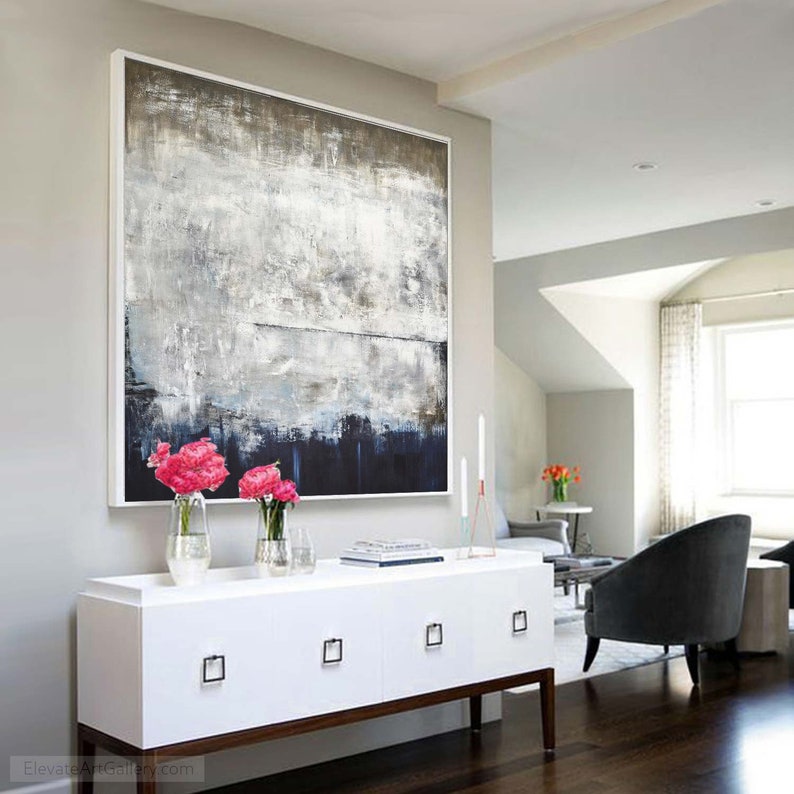 May include: A white dresser with four drawers and black square handles. The dresser is in front of a large abstract painting with shades of grey, white, and blue. There are two vases with pink flowers on top of the dresser.