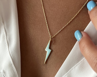 May include: A gold-colored necklace with a lightning bolt pendant. The lightning bolt is light blue and gold. The necklace is worn over a white shirt.