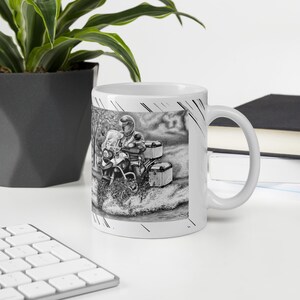 May include: A white ceramic mug with a black and white illustration of a person riding a motorcycle on a dirt road. The motorcycle has luggage attached to the back.