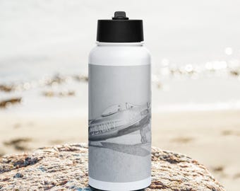 May include: A white water bottle with a black lid and a black and white photo of a vintage airplane with the text "US NAVY" on the side. The bottle is sitting on a rock.