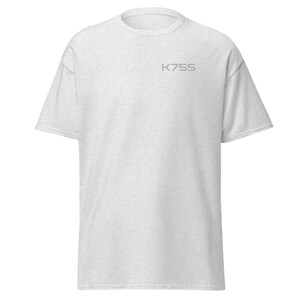 以下が含まれることがあります： クルーネックのライトヘザーグレーの半袖Tシャツ。左胸上部にK75Sと書かれた小さなダークグレーのテキストグラフィックがプリントされています。標準的なフィット感で、裾はストレートなシンプルなデザインの衣類です。