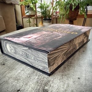 Puede incluir: Un libro de tapa dura titulado "Secrets of the Alchemised" con una cubierta oscura que presenta ilustraciones arquitectónicas. Los bordes del libro tienen diseños detallados en relieve. El libro está sobre una superficie texturizada.