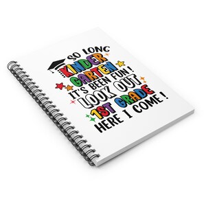 May include: A white spiral notebook with the text "SO LONG KINDER GARDEN IT'S BEEN FUN! LOOK OUT 1ST GRADE HERE I COME!" in colorful letters. The text is decorated with stars and a graduation cap.