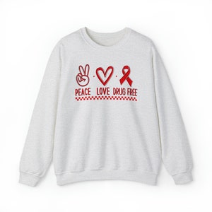May include: A light gray sweatshirt with a red peace sign, heart, and ribbon. The text "Peace Love Drug Free" is printed below the symbols.