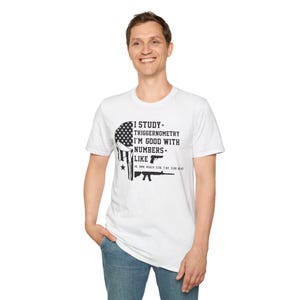 May include: White t-shirt with a graphic design featuring a skull with an American flag pattern and the text "I STUDY TRIGGERNOMENTRY I'M GOOD WITH NUMBERS LIKE" with gun illustrations. The shirt is worn by a person.