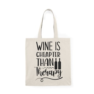 May include: A white canvas tote bag with black text that reads "Wine is cheaper than therapy" with three wine bottle silhouettes.