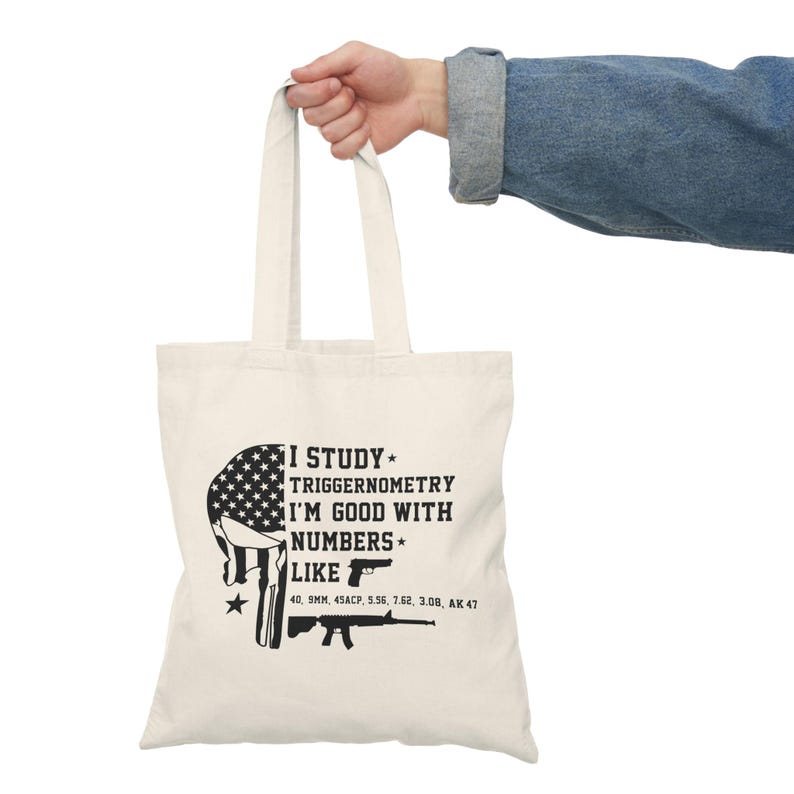 May include: Off-white canvas tote bag with black text and graphics. The text reads "I STUDY TRIGGERNOMETRY I'M GOOD WITH NUMBERS LIKE" followed by a list of gun calibers. The graphic is a skull with the American flag.