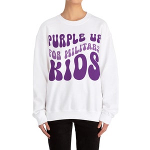 Puede incluir: Sudadera blanca con un gr&aacute;fico morado que dice "Purple Up For Military Kids" en una fuente retro.