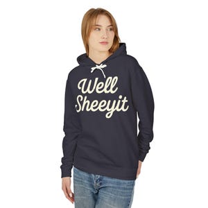 K&ouml;nnte beinhalten: Ein marineblauer Kapuzenpullover mit dem cremefarbenen Text "Well Sheeyit" in einer Schriftart im Vintage-Stil. Der Hoodie hat einen Kordelzug und eine Vordertasche. Das Model tr&auml;gt eine blaue Jeans.
