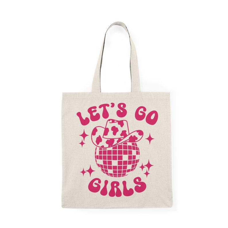 May include: A white tote bag with a pink design. The design features a disco ball with a pink cowboy hat on top. The text "Let's Go Girls" is written in pink.
