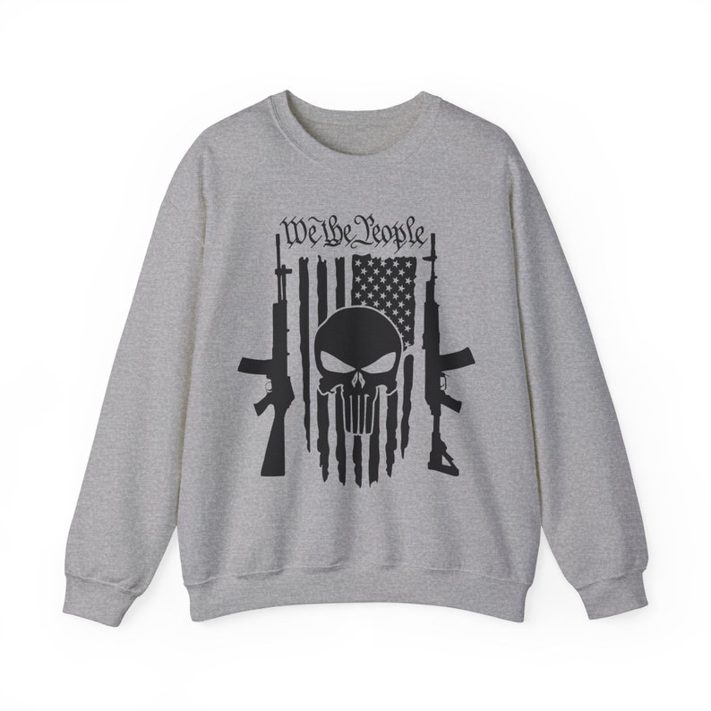 Op de afbeelding: Een grijs sweatshirt met een zwarte schedel en gekruiste beenderen afbeelding. De schedel bevindt zich voor een Amerikaanse vlag met de woorden "We the People" erboven.