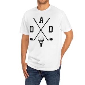 May include: White t-shirt with a black graphic design featuring the word "DAD" with crossed golf clubs, a golf ball, and a tee. The shirt is made of a soft material and is suitable for casual wear.