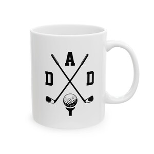 May include: White ceramic mug with a black graphic design. The design features the word "DAD" in bold letters, with crossed golf clubs, a golf ball, and a tee. The mug has a curved handle.