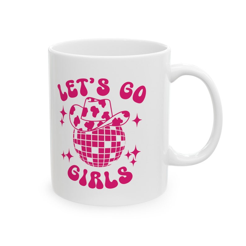 May include: White ceramic mug with a pink design. The design features a disco ball with a cow print cowboy hat on top. The text "Let's Go Girls" is written in pink.