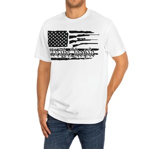 Op de afbeelding: Wit T-shirt met een zwart-wit ontwerp van een verweerde Amerikaanse vlag. De vlag heeft een zwart silhouet van geweren en de tekst "We the People" in een gestileerd lettertype.