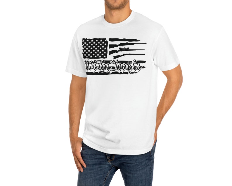 Op de afbeelding: Wit T-shirt met een zwart-wit ontwerp van een verweerde Amerikaanse vlag. De vlag heeft een zwart silhouet van geweren en de tekst "We the People" in een gestileerd lettertype.