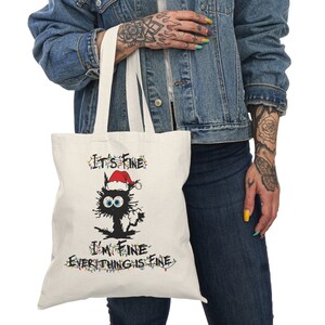 May include: White canvas tote bag with a black cat wearing a Santa hat and surrounded by Christmas lights. The text on the bag reads "It's Fine I'm Fine Everything is Fine."