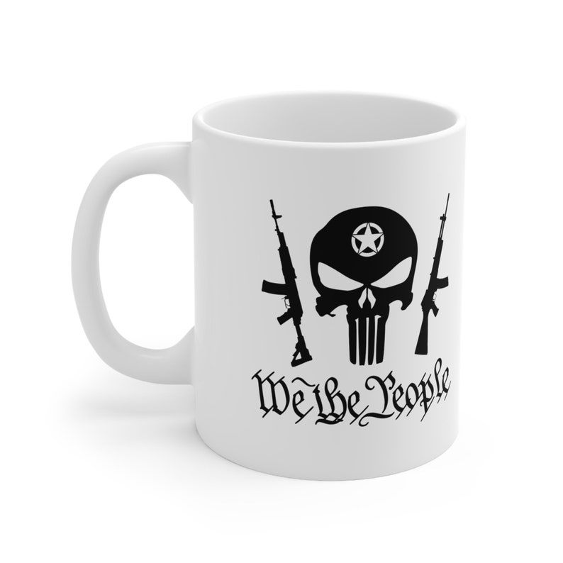 Op de afbeelding: Witte keramische koffiemok met een zwart silhouet van een schedel met een ster op zijn voorhoofd, twee geweren en de tekst "We the People".