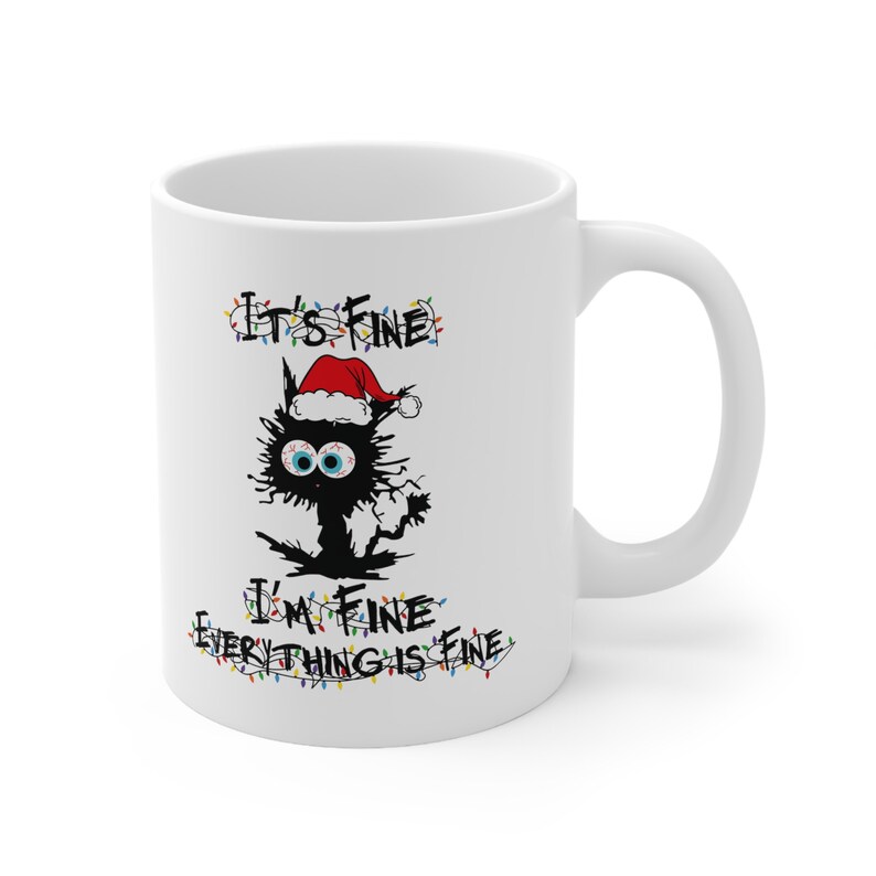 May include: White ceramic mug with a black cat wearing a Santa hat and Christmas lights. The text on the mug reads "It's Fine I'm Fine Everything is Fine."