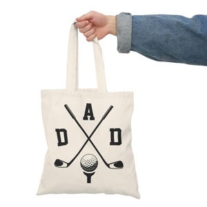 May include: Cream-colored tote bag with the word "DAD" in black block letters, with crossed golf clubs and a golf ball graphic. The bag is held by a person wearing a denim jacket.