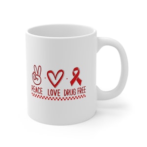 May include: White ceramic coffee mug with a red peace sign, heart, and ribbon with the text "Peace Love Drug Free" and a red and white checkered border.
