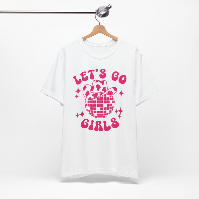 May include: White t-shirt with a pink graphic of a disco ball wearing a cowboy hat with a cow print design. The text "Let's Go Girls" is printed in pink.