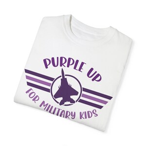 Puede incluir: Camiseta blanca con un dise&ntilde;o morado que dice "Purple Up For Military Kids". El dise&ntilde;o presenta una silueta morada de un avi&oacute;n de combate con alas y rayas.