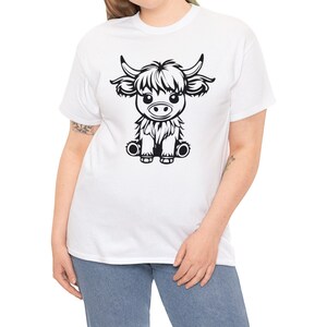 以下が含まれることがあります： 白いTシャツに、座っているハイランド牛の白黒のイラストが描かれています。牛はふわふわのたてがみと可愛い表情をしています。