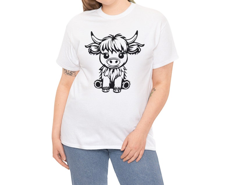 以下が含まれることがあります： 白いTシャツに、座っているハイランド牛の白黒のイラストが描かれています。牛はふわふわのたてがみと可愛い表情をしています。
