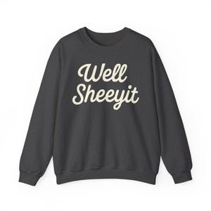 K&ouml;nnte beinhalten: Dunkelgraues Sweatshirt mit dem Schriftzug "Well Sheeyit" in wei&szlig;er, kursiver Schrift. Das Sweatshirt hat einen Rundhalsausschnitt und lange &Auml;rmel. Der Text ist mittig auf der Brust platziert.