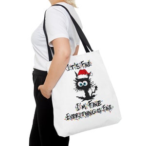 May include: White tote bag with a black cat wearing a Santa hat and surrounded by Christmas lights. The text on the bag reads "It's Fine I'm Fine Everything is Fine."