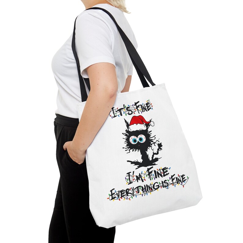 May include: White tote bag with a black cat wearing a Santa hat and surrounded by Christmas lights. The text on the bag reads "It's Fine I'm Fine Everything is Fine."