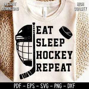 May include: A cream-coloured t-shirt featuring a black graphic design of a hockey helmet, a hockey stick, and a puck. The text reads EAT SLEEP HOCKEY REPEAT in a bold, black, block-style font. The design is centred on the front of the shirt.