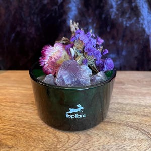 May include: A dark green glass bowl featuring a white graphic of a running rabbit and the text Hop Hare. The bowl is filled with rough purple amethyst crystals, a single pink strawflower, and sprigs of dried purple statice flowers.
