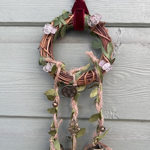 May include: A brown twig wreath with green leaves and clear crystal accents hangs from a burgundy ribbon. Three bells hang from the wreath, each with a key charm and green leaves.