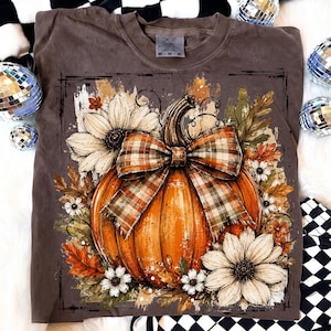 May include: A brown Comfort Colors t-shirt featuring a vibrant orange pumpkin with a plaid bow, surrounded by white and orange flowers and autumn leaves. The design evokes a fall or harvest theme.