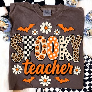 K&ouml;nnte beinhalten: Ein braunes Kurzarm-T-Shirt mit dem Schriftzug "SPOOKY teacher" in gro&szlig;en, gemusterten Buchstaben. Das Design zeigt orangefarbene Flederm&auml;use, wei&szlig;e G&auml;nsebl&uuml;mchen, Leopardenmuster, schwarz-wei&szlig;e Karos, K&uuml;rbisse, Punkte und Sterne.