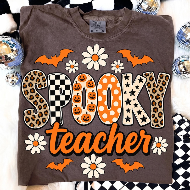K&ouml;nnte beinhalten: Ein braunes Kurzarm-T-Shirt mit dem Schriftzug "SPOOKY teacher" in gro&szlig;en, gemusterten Buchstaben. Das Design zeigt orangefarbene Flederm&auml;use, wei&szlig;e G&auml;nsebl&uuml;mchen, Leopardenmuster, schwarz-wei&szlig;e Karos, K&uuml;rbisse, Punkte und Sterne.