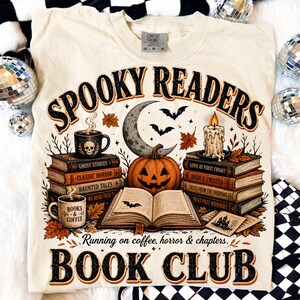 May include: A cream-colored t-shirt featuring a graphic print with the text "SPOOKY READERS" and "RUNNING ON COFFEE, HORROR & CHAPTERS, BOOK CLUB". The design includes stacks of books, a jack-o'-lantern, a crescent moon, bats, and a candle.
