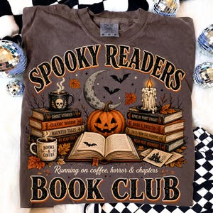 May include: A brown t-shirt featuring a graphic with the text SPOOKY READERS and BOOK CLUB. The design includes stacks of books, a jack-o'-lantern, a crescent moon, bats, a candle, and two mugs with the text BOOKS & COFFEE and skull imagery.